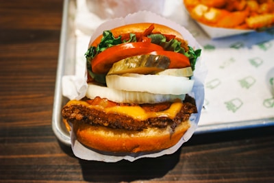 A close-up of a cheeseburger featuring crispy fried patty, melted cheese, fresh lettuce, slices of tomato, pickles, and onion between a soft bun. The burger is served on a rectangular metal tray, with a side of crinkle-cut fries visible in the background.