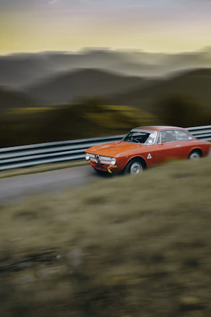 A sepia-toned vintage racing car speeding along a winding mountain road under a cloudy sky.