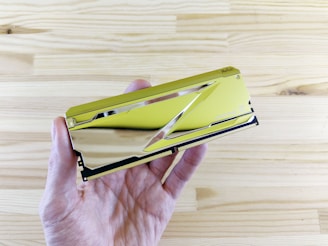 A close-up of a hand holding a gold-colored computer RAM module against a light wooden background. The RAM's surface is reflective, creating a sleek and futuristic appearance.