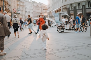 A busy urban street scene with a mix of pedestrians walking in different directions. An adorable child in white clothes stands in the foreground, surrounded by adults. The environment seems lively with a variety of people, some casually dressed, including a man in a blue shirt and a woman with a floral top. A bicycle rickshaw is visible in the background, and the architectural backdrop suggests a bustling city atmosphere with sunlight illuminating the scene.