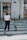 A person in a white shirt and black pants is walking a small brown dog on a leash across a zebra crossing in an urban setting. A white building with a sign and graffiti is in the background.