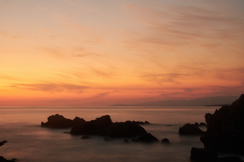 the sun is setting over the ocean with rocks in the foreground