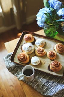 A wooden table holds a baking tray with freshly baked cinnamon rolls, some of which are topped with icing. Beside the tray is a white coffee mug filled with dark coffee. A textured gray cloth is placed underneath. In the background, a green vase holds a bouquet of vibrant blue hydrangeas.