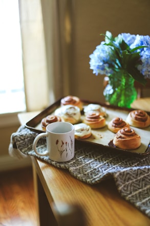 A vibrant table spread with cinnamon rolls, mini pizzas, and steaming coffee cups bathed in warm terracotta light.