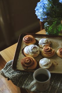 A tray of freshly baked cinnamon rolls, some topped with icing, sits on a wooden table next to a patterned cloth. A cup of coffee is placed in front of the tray, and a vibrant green glass vase holds a bouquet of blue hydrangeas beside it.