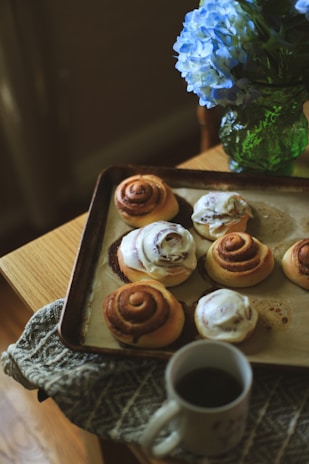 A tray of freshly baked cinnamon rolls, some topped with icing, sits on a wooden table next to a patterned cloth. A cup of coffee is placed in front of the tray, and a vibrant green glass vase holds a bouquet of blue hydrangeas beside it.