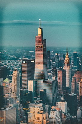 The image depicts a cityscape with a focus on a modern skyscraper prominently positioned in the foreground. Surrounding it are various other high-rise buildings, showcasing diverse architectural styles. The sky above is clear, and the city stretches out into the distance, implying a bustling metropolis.