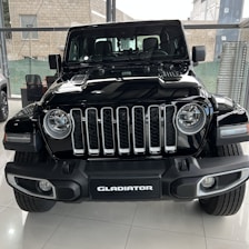 A black Jeep Gladiator is parked indoors in a well-lit showroom. It features a prominent front grille with seven vertical slots, rounded headlights, and sturdy bumper design. In the background, other vehicles and showroom elements like glass walls and a seating area are visible.