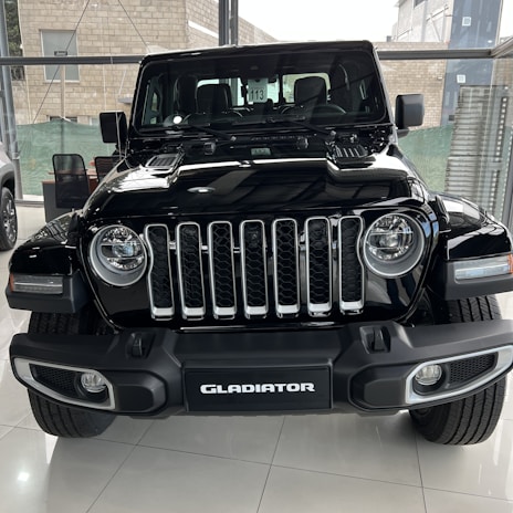A black Jeep Gladiator is parked indoors in a well-lit showroom. It features a prominent front grille with seven vertical slots, rounded headlights, and sturdy bumper design. In the background, other vehicles and showroom elements like glass walls and a seating area are visible.