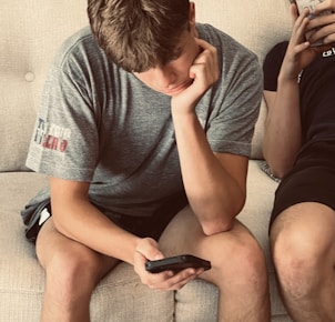 a man sitting on a couch next to another man holding a cell phone