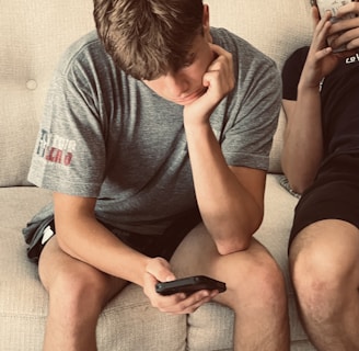 a man sitting on a couch next to another man holding a cell phone