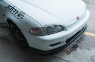 The image features the front part of a white car with noticeable water droplets on its surface. The car has a Honda emblem and Japanese characters near the headlight. It also sports decals on the side indicating JDM and other graphics. The vehicle is parked on a wet concrete surface.