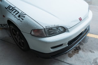 The image features the front part of a white car with noticeable water droplets on its surface. The car has a Honda emblem and Japanese characters near the headlight. It also sports decals on the side indicating JDM and other graphics. The vehicle is parked on a wet concrete surface.