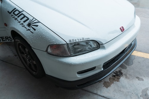 The image features the front part of a white car with noticeable water droplets on its surface. The car has a Honda emblem and Japanese characters near the headlight. It also sports decals on the side indicating JDM and other graphics. The vehicle is parked on a wet concrete surface.