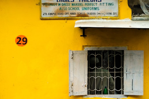 A bright yellow wall features a vintage tailor shop sign above a window with bars and partially opened shutters. The number 29 is prominently displayed on the wall in a red circle. Behind the window, a green bottle is visible along with a faint outline of a person.