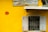 A bright yellow wall features a vintage tailor shop sign above a window with bars and partially opened shutters. The number 29 is prominently displayed on the wall in a red circle. Behind the window, a green bottle is visible along with a faint outline of a person.