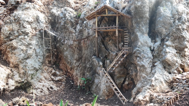 A rocky cliffside with a small wooden structure perched against the rock. The structure consists of a platform accessible via a narrow wooden ladder and a spiral staircase. A simple rope bridge connects two parts of the cliff. Sparse vegetation grows at the base of the cliff.