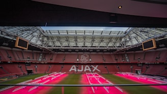 A large football stadium with a prominent logo in the center of the stands, showcasing the name 'AJAX' on red seats. The stadium's roof is partially open, and there are electronic screens on either side. The playing field features artificial lighting equipment, casting a pinkish glow over the grass.