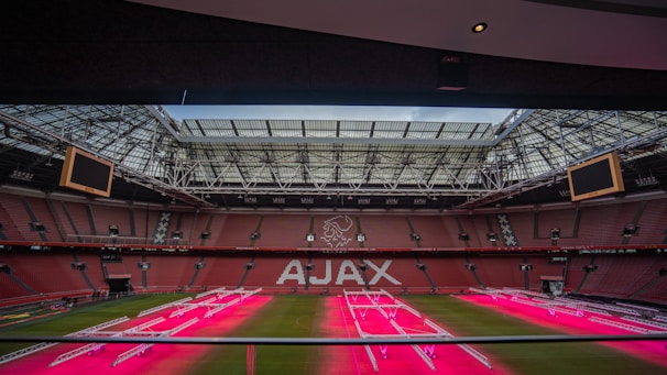 A large football stadium with a prominent logo in the center of the stands, showcasing the name 'AJAX' on red seats. The stadium's roof is partially open, and there are electronic screens on either side. The playing field features artificial lighting equipment, casting a pinkish glow over the grass.