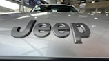 Close-up view of a car hood with the word 'Jeep' prominently displayed in dark, bold letters. The background shows an indoor car showroom with other vehicles and showroom decor.