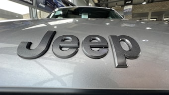 Close-up view of a car hood with the word 'Jeep' prominently displayed in dark, bold letters. The background shows an indoor car showroom with other vehicles and showroom decor.