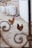 Three chickens roam around a courtyard area, featuring stone pavement and a brick wall partially covered by a white textured wall. Large white bags are stacked in the background, suggesting a storage or construction setting. The view is partially framed by intricate metal scrollwork.