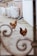 Three chickens roam around a courtyard area, featuring stone pavement and a brick wall partially covered by a white textured wall. Large white bags are stacked in the background, suggesting a storage or construction setting. The view is partially framed by intricate metal scrollwork.