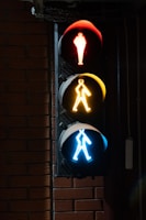 A vertical traffic light with three distinct lights arranged top to bottom. The top light is red with a standing figure, the middle light is yellow with a figure walking, and the bottom light is blue with a figure in motion. The traffic light is mounted on a brick wall and is illuminated against the dark background.