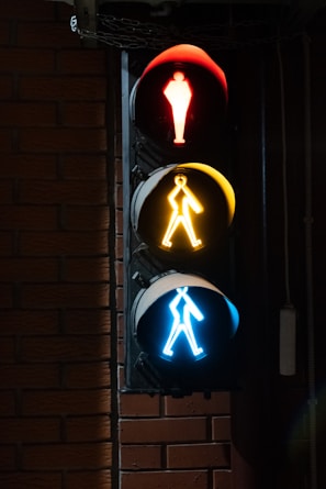 A vertical traffic light with three distinct lights arranged top to bottom. The top light is red with a standing figure, the middle light is yellow with a figure walking, and the bottom light is blue with a figure in motion. The traffic light is mounted on a brick wall and is illuminated against the dark background.