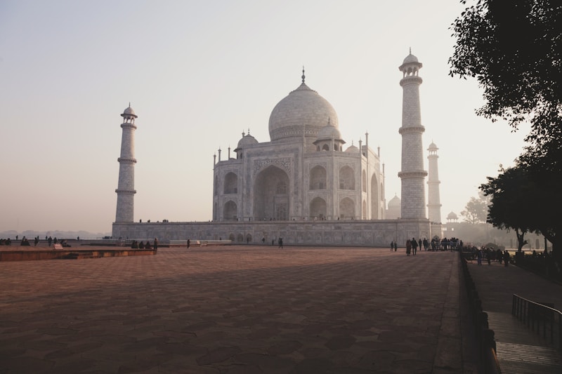 The image captures a grand architectural monument made of white marble, featuring a large dome surrounded by four minarets. There is a spacious courtyard in the foreground, and the scene is enveloped in soft, ambient lighting with a slight haze, creating an ethereal atmosphere. Visitors can be seen walking and gathering around the structure.