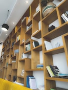 A wooden bookshelf spans an entire wall, filled with various books and scattered decorative items. Some of the shelves hold plants and woven baskets, while books of different sizes and colors are arranged both vertically and horizontally. The shelves create an intricate geometric pattern, with some sections empty or sparsely filled. Overhead lighting adds illumination to the scene.