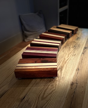 Rectangular wooden blocks are arranged diagonally on a wooden table. Each block features a polished finish with contrasting light and dark wood stripes. The table has a visible grain pattern and there is a cushion in the background.