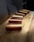 Rectangular wooden blocks are arranged diagonally on a wooden table. Each block features a polished finish with contrasting light and dark wood stripes. The table has a visible grain pattern and there is a cushion in the background.