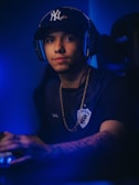 A person wearing a cap and headphones sits in a gaming chair, looking at the viewer with a neutral expression. They are wearing a shirt with a sports team logo and a gold chain. The lighting is predominantly blue, creating a calm atmosphere.