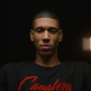 A portrait of a young man with a neutral expression, wearing a black shirt with red script against a dark background. The lighting is focused on his face, creating an intimate atmosphere.