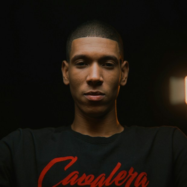 A portrait of a young man with a neutral expression, wearing a black shirt with red script against a dark background. The lighting is focused on his face, creating an intimate atmosphere.
