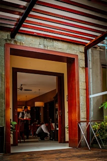 A restaurant or café interior with warm lighting and wooden accents. Two people are present, one standing with a tray and another crouching, seemingly adjusting something. The setting has chairs, tables, potted plants, and hanging ceiling lamps.