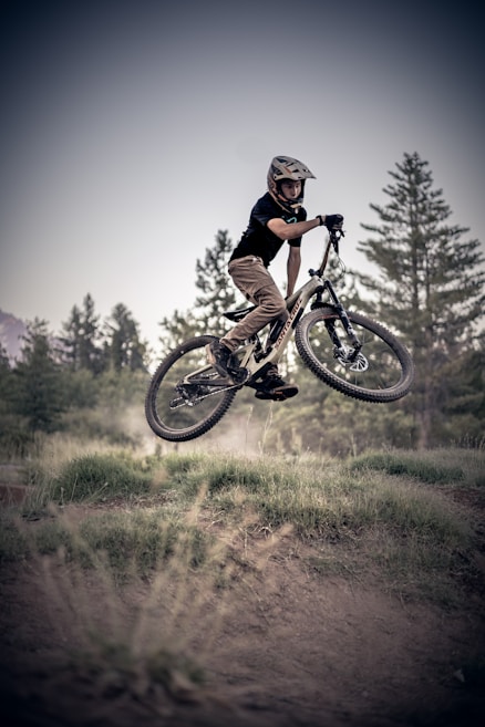 A cyclist performs a jump on a mountain bike, wearing protective gear including a helmet. The background features tall pine trees and a clear sky, suggesting an outdoor setting suitable for biking.