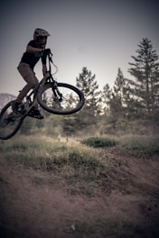 A person wearing a helmet is performing a jump on a mountain bike in an outdoor setting. The background features a line of trees and an overcast sky, and the scene is captured mid-action, emphasizing the thrill of mountain biking.