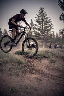 A person is riding a mountain bike over a small dirt mound in a forested area. The background includes tall trees and blurred figures in the distance, suggesting a natural setting suitable for biking. The rider wears a helmet and casual athletic clothing.