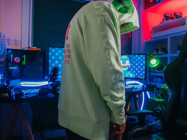 A person wearing a light green hoodie stands in a room equipped with a gaming setup. The setup includes multiple monitors with one displaying a character or game. There is a vibrant neon lighting effect present in the room, creating a colorful atmosphere. Shelving with shoes can be seen in the background.
