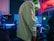 A person wearing a light green hoodie stands in a room equipped with a gaming setup. The setup includes multiple monitors with one displaying a character or game. There is a vibrant neon lighting effect present in the room, creating a colorful atmosphere. Shelving with shoes can be seen in the background.