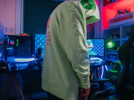 A person wearing a light green hoodie stands in a room equipped with a gaming setup. The setup includes multiple monitors with one displaying a character or game. There is a vibrant neon lighting effect present in the room, creating a colorful atmosphere. Shelving with shoes can be seen in the background.