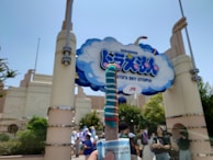 An amusement park entrance featuring a large sign with cloud and cartoon graphics, held by ornate pillars. People are gathered below, some taking photos, while a vibrant blue and red churro is prominently held in the foreground.