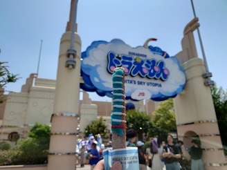 An amusement park entrance featuring a large sign with cloud and cartoon graphics, held by ornate pillars. People are gathered below, some taking photos, while a vibrant blue and red churro is prominently held in the foreground.