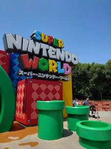 A colorful sign with the words 'Super Nintendo World' stands prominently, surrounded by large green pipes reminiscent of a video game landscape. Bright blocks with patterns enhance the scene, set against a clear blue sky. People, including a person pushing a stroller, are nearby, suggesting a theme park atmosphere.