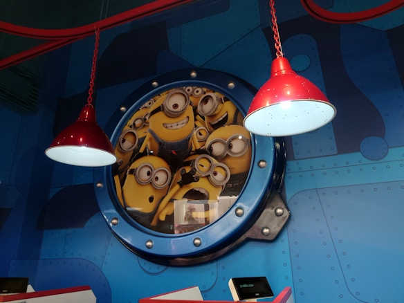 A group of animated yellow characters with goggles are peering through a circular window against a backdrop resembling a submarine's interior. Two red hanging lamps are prominently displayed above them, casting light on the characters.