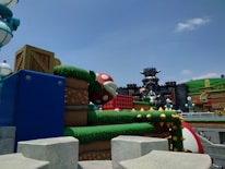 An outdoor theme park scene featuring colorful, cartoon-style architecture. Prominent elements include a large, red mushroom with white spots, blocks resembling those from a video game, a castle with horned features, and surrounding green hills. The overall aesthetic is whimsical and fantastical.