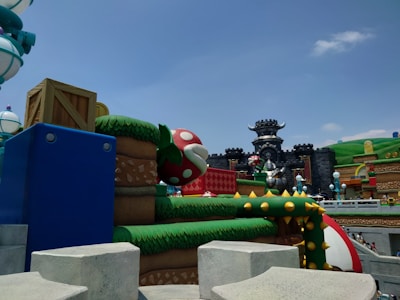 An outdoor theme park scene featuring colorful, cartoon-style architecture. Prominent elements include a large, red mushroom with white spots, blocks resembling those from a video game, a castle with horned features, and surrounding green hills. The overall aesthetic is whimsical and fantastical.