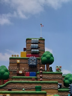 A themed amusement park area designed to resemble a level from a video game, featuring hills and platforms made of bricks and grass. Various iconic elements from the game series, such as Goombas, Thwomps, and blocks with coins, are scattered throughout the scene. Artificial trees add to the colorful and playful landscape.